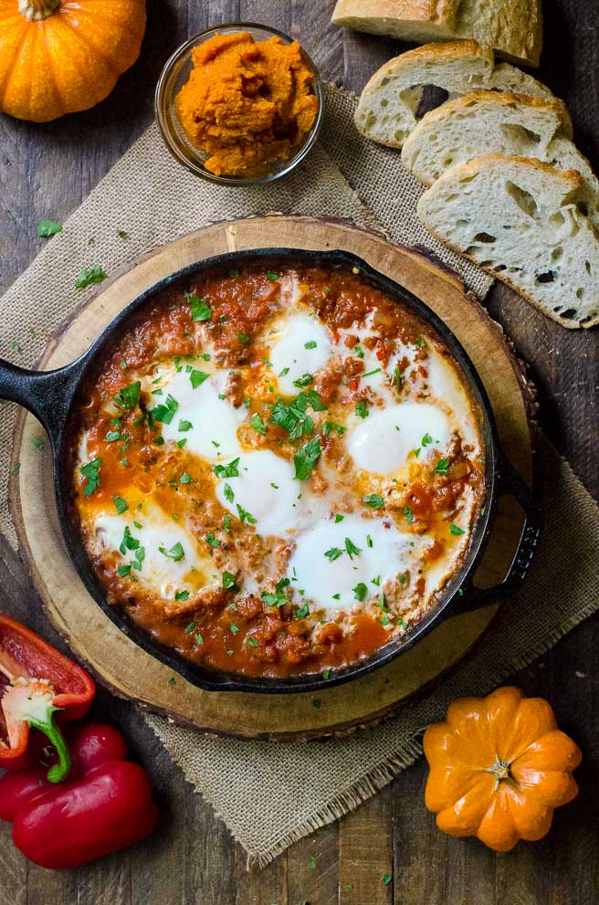 Overhead view of a wooden table with a pan of pumpkin shakshuka in the middle.  The table is decorated for fall.