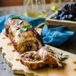 A table set with sliced stuffed pork tenderloin next to glasses of white wine and a box of prunes.