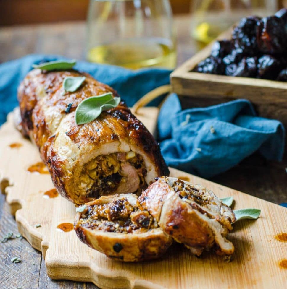 A table set with sliced stuffed pork tenderloin next to glasses of white wine and a box of prunes.