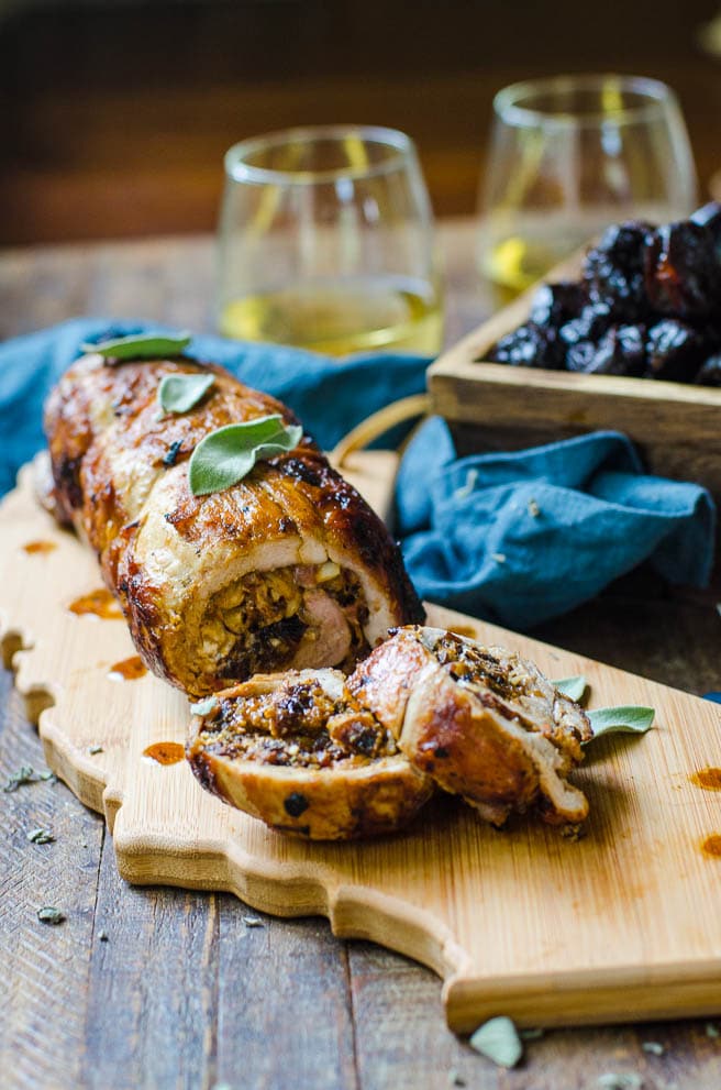 A table set for serving a cooked stuffed pork tenderloin.  It is surrounded by a box of prunes and white wine in glasses.