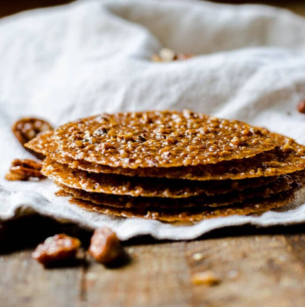 A stack of pecan lace cookies surrounded by candied pecans.