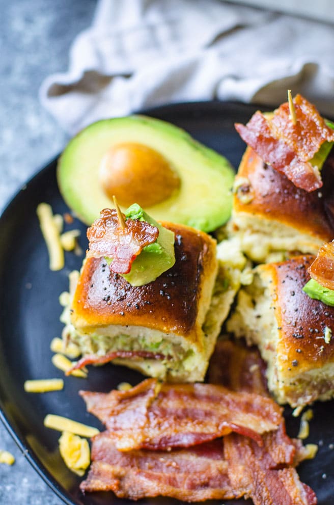 A black plate filled hawaiian roll turkey sliders, cooked bacon, and avocado.