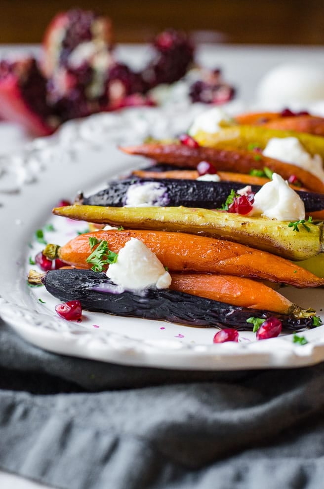 A white platter of roasted rainbow carrots garnished with burrata and pomegranate seeds.