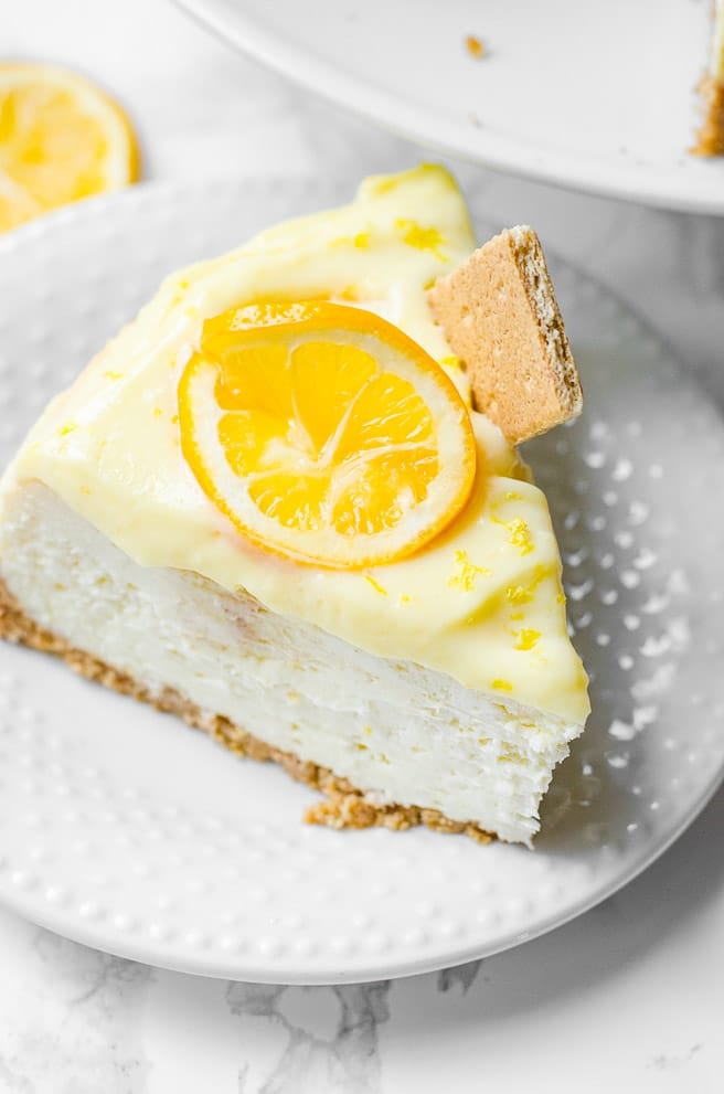 A single slice of lemon cheesecake on a plate. It is garnished with a lemon wheel and graham cracker.