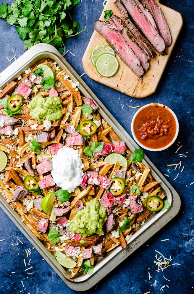 Overhead view of a sheet pan of carne asada fries next to a cutting board of carne asada steak ready for serving.