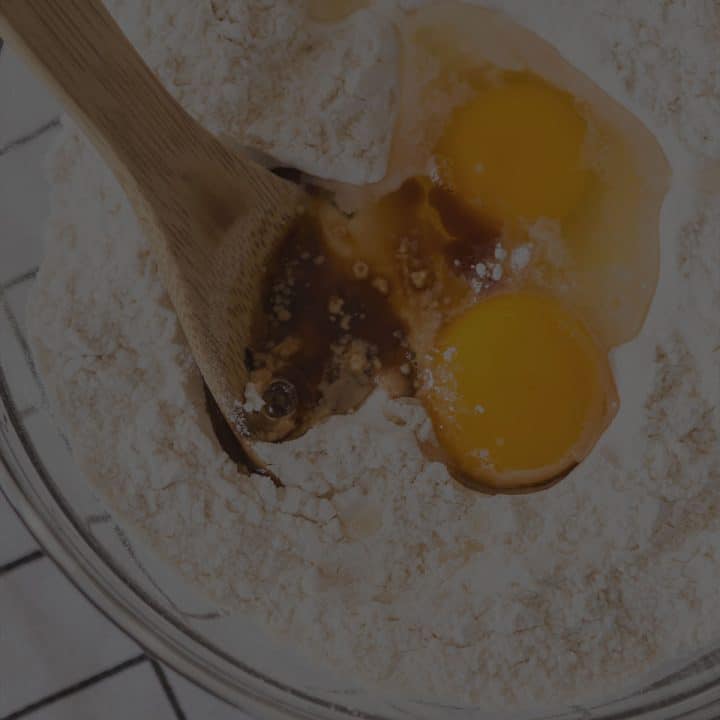A wooden spoon getting ready to stir cracked eggs into a bowl of flour.