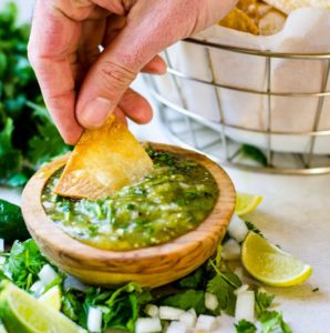 A hand dipping a tortilla chip into tomatillo salsa.