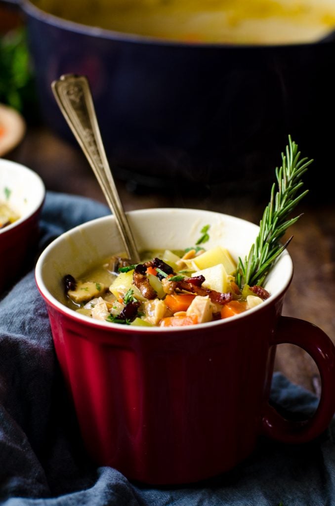 A red soup mug of soup with a spoon and rosemary garnish.