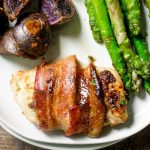 Bacon wrapped chicken thigh on a plate next to roasted potatoes and asparagus.