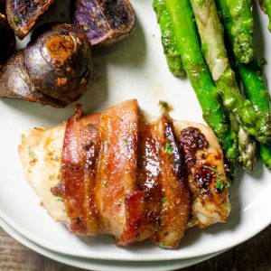 Bacon wrapped chicken thigh on a plate next to roasted potatoes and asparagus.