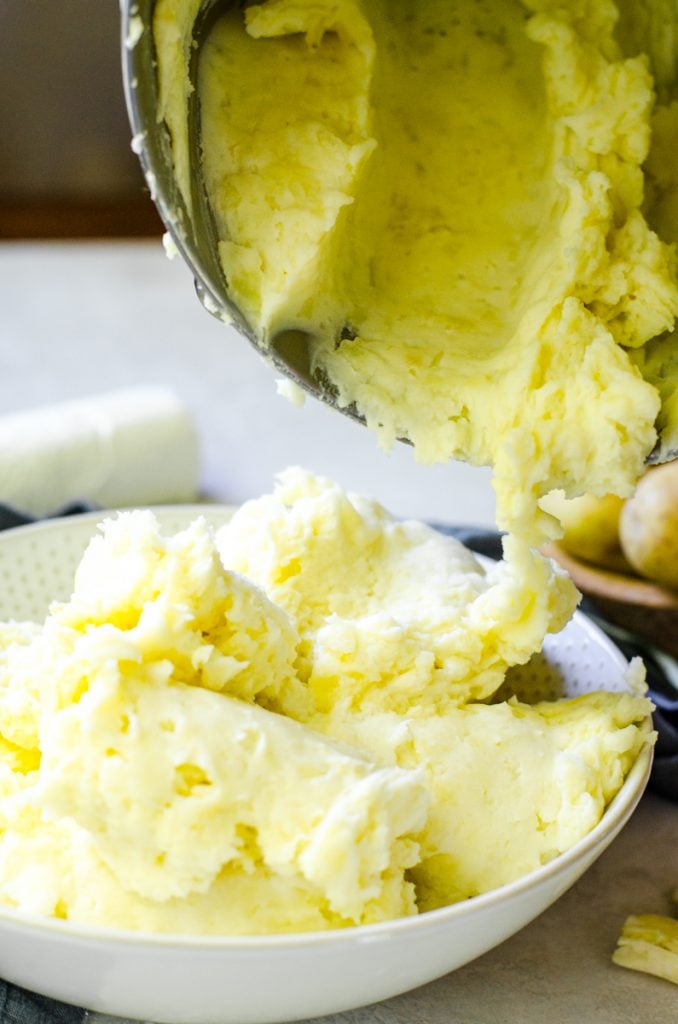 Goat cheese mashed potatoes being scraped out of a mixing bowl and into a serving bowl.