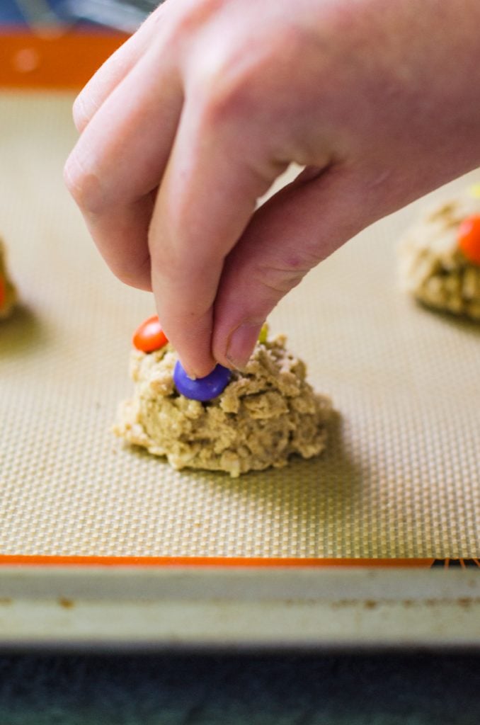 A hand pushing a purple M&M into cookie dough.