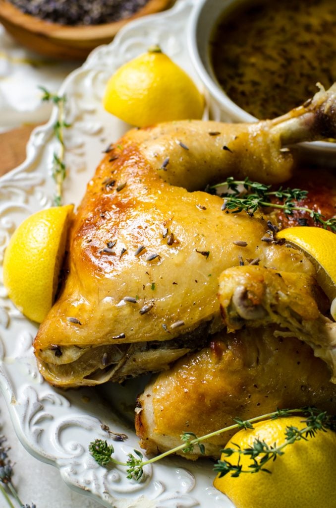 A platter of pan juices in a bowl next to pieces of lemon lavender chicken garnished with lemon wedges and fresh thyme.