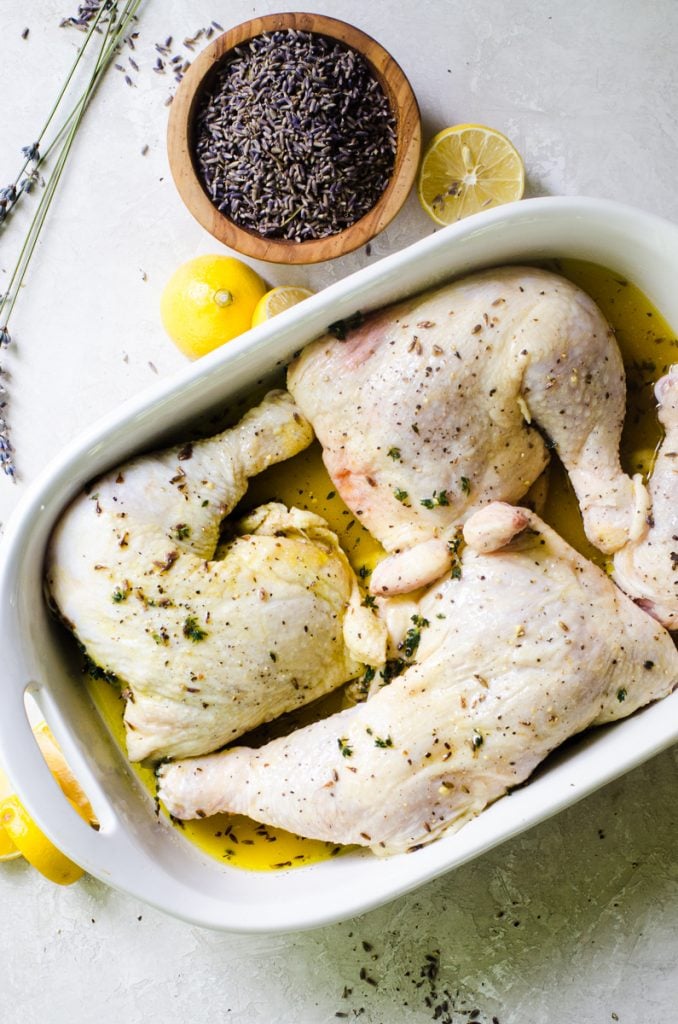 Overhead view of chicken leg quarters in a baking dish next to a bowl of lavender and lemon wedges.