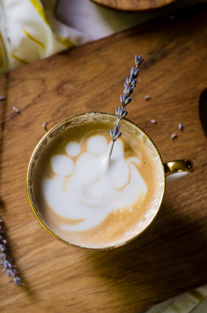 A stem of lavender sticking out of a latte on a wooden board.