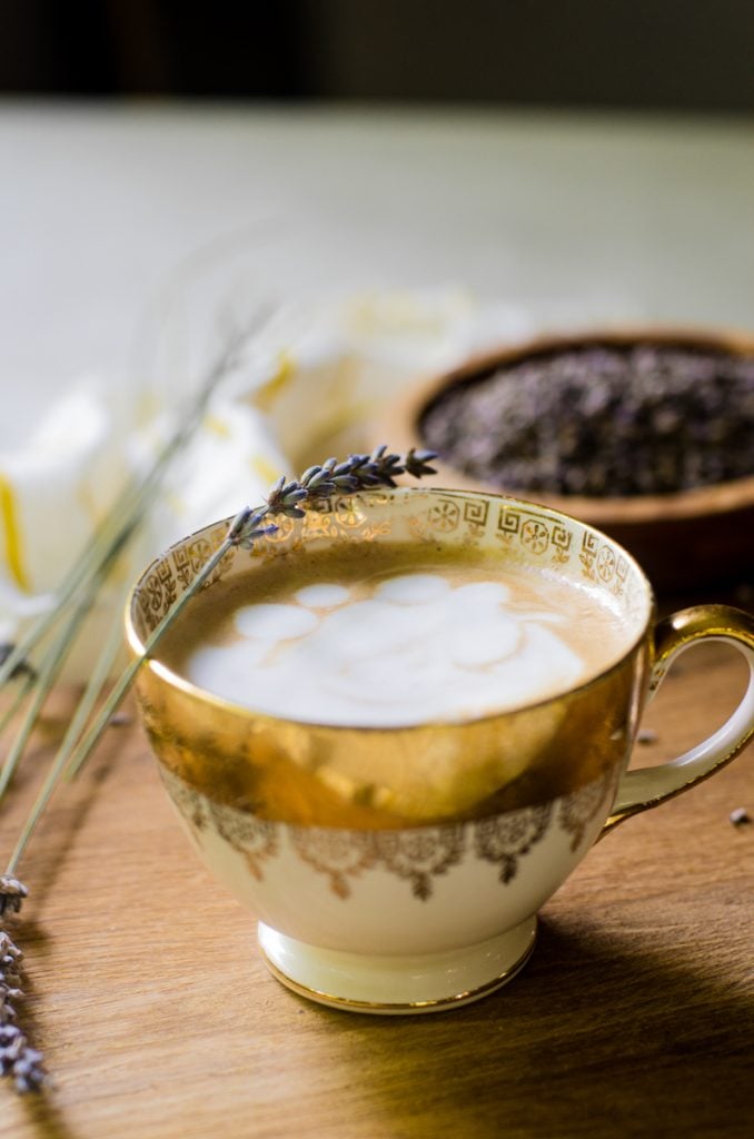 A wooden board with a lavender latte in a golden cup on it.  It is garnished with a stem of fresh lavender.