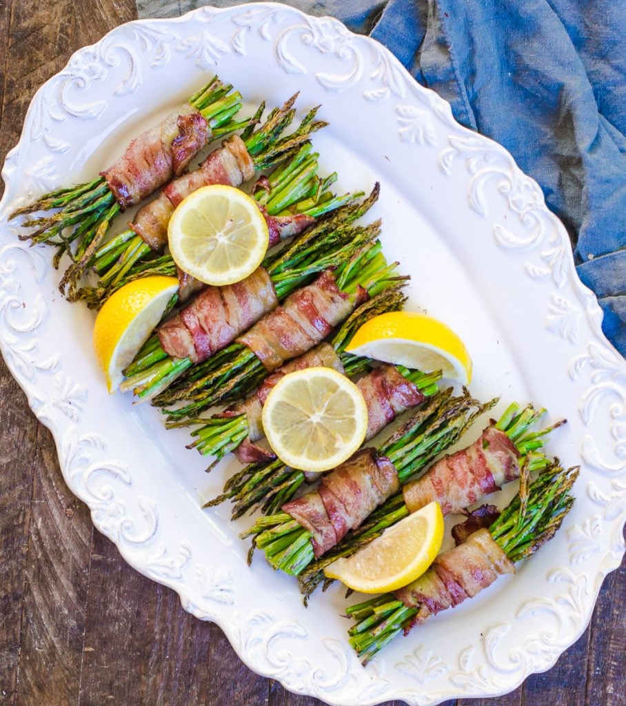 Bundles of asparagus on a white plate garnished with lemon wheels and wedges.