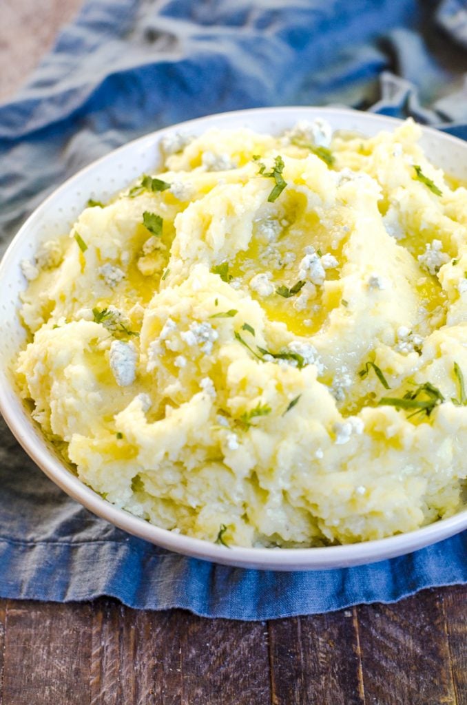 A bowl of blue cheese mashed potatoes garnished with blue cheese and butter on a blue napkin.