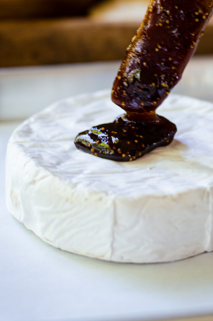 A wooden spreading knife putting fig jam on a wheel of brie.