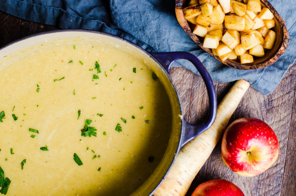 A dutch oven of soup next to a wooden bowl of apples. Parsnips and apples are next to them.