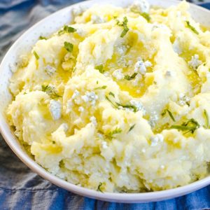 A large bowl of mashed potatoes garnished with blue cheese and melted butter.
