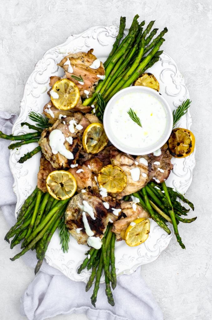 An overhead view of a white tray of grilled chicken and asparagus next to grilled lemons and yogurt sauce.