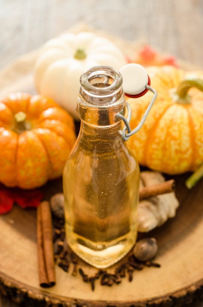 A jar of pumpkin pie spice syrup on a wooden board in front of decorative pumpkins.