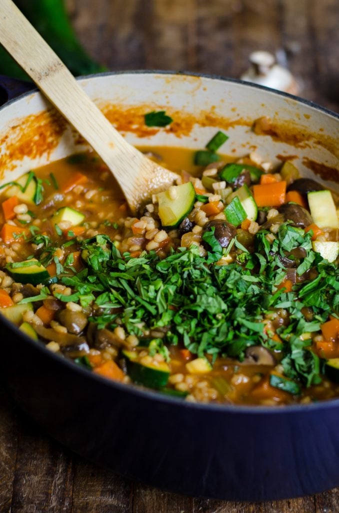 A purple pot of vegetable barley soup with green garnish and a wooden spoon sticking out of it.