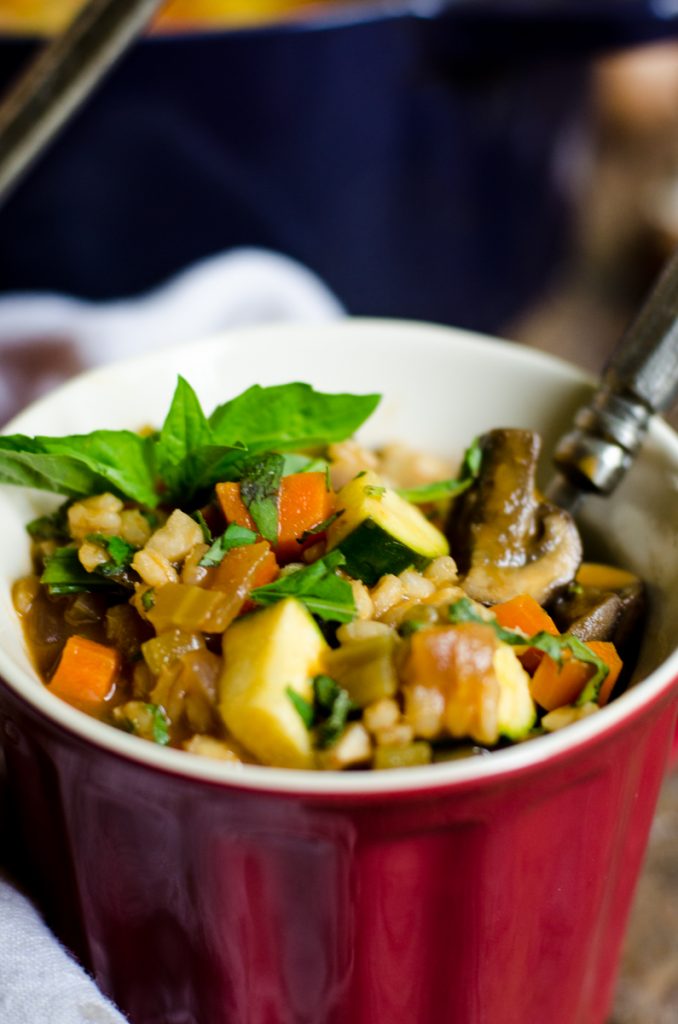 Close up of vegetable barley soup inside of a red mug with a spoon sticking out.