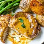 A cut pork chop with sauce next to green beans and potatoes.
