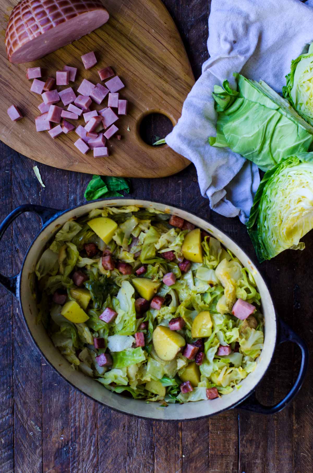 Overhead flatlay of a pot of ham and cabbage next to a head of cabbage and cutting board of ham.