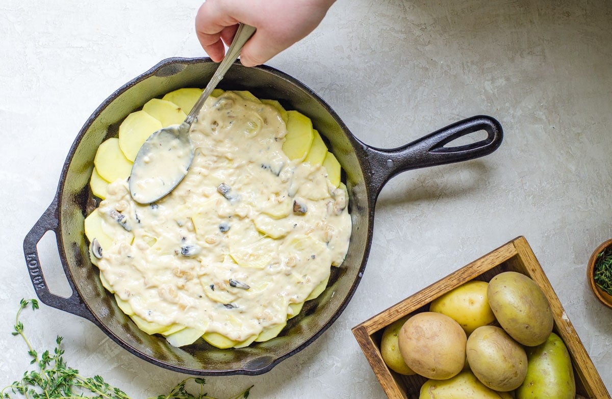 A hand spreading out cream of mushroom cheese sauce on Scalloped Potatoes with Mushroom Soup