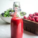 A bottle of raspberry vinaigrette dressing in front of a salad and raspberries.