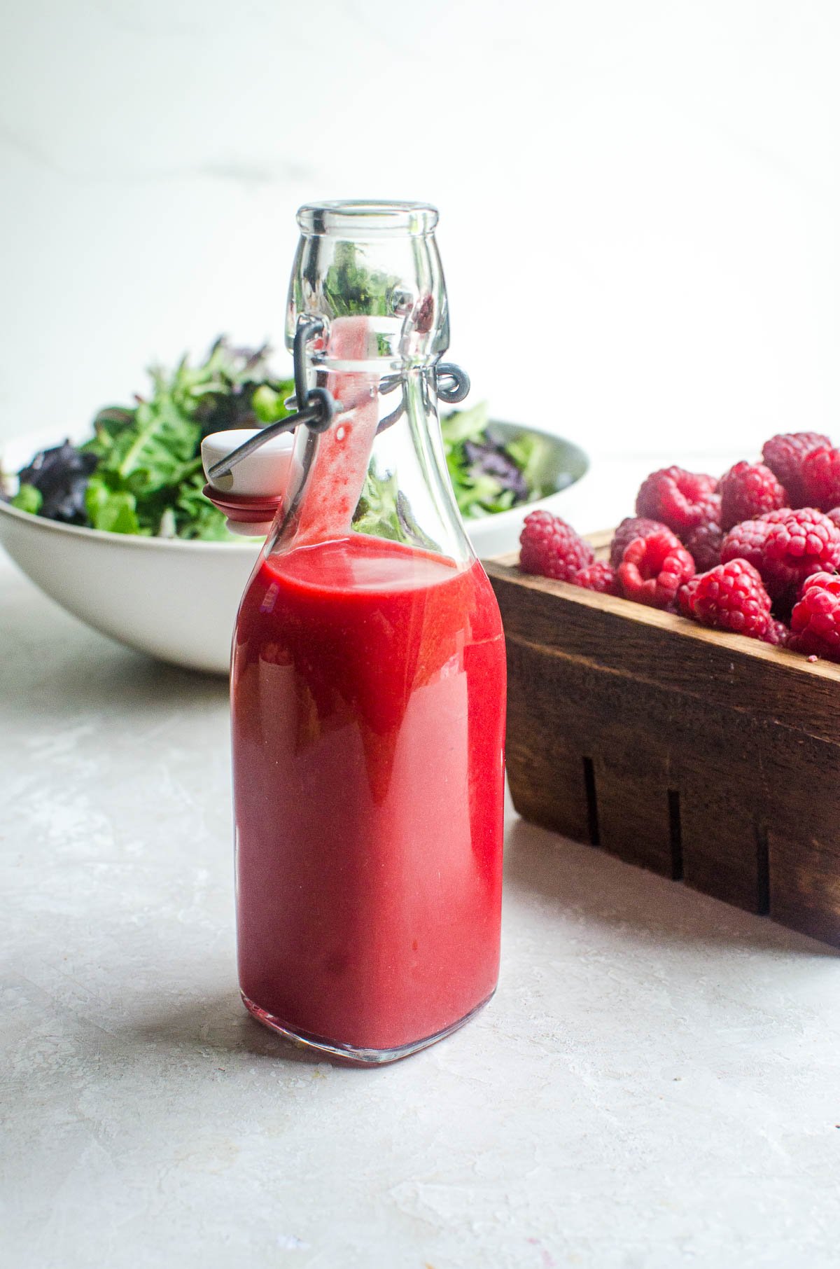 A bottle of raspberry vinaigrette dressing in front of a box of raspberries and bowl of salad.