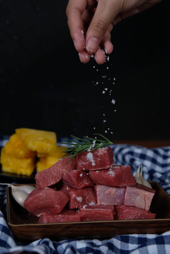 A hand showing how to season food by sprinkling salt on cubed meat.